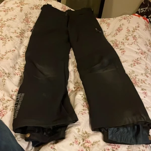 Svarta täckbyxor från Windy Peak - Svarta täckbyxor från Windy Peak North Team Expedition med elastiska benslut och tryckknapp. Byxorna har dragkedja och är fodrade för extra värme, perfekta för kalla dagar. Snygg logga på benet och praktiska fickor med dragkedja.Tillhörande hängslen finns.