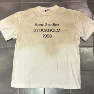 Beige Acne Studios t-shirt Stockholm - Säljer en beige t-shirt från Acne Studios med svart text 'STOCKHOLM 1996' på bröstet. T-shirten har en cool gradient-effekt och klassisk rund hals. Materialet är mjuk bomull och modellen är normal passform med korta ärmar.