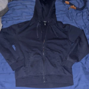 Mörkblå hoodie med dragkedja och huva - Mörkblå hoodie med hel dragkedja framtill och huva med snörning. Tröjan har långa ärmar, ribbade muddar och två fickor på framsidan. Perfekt för chill dagar eller när du vill ha en enkel och stilren look.