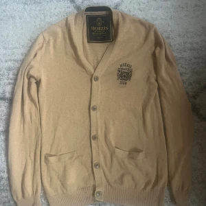 Beige kofta från Morris Ivy League - Snygg beige kofta från Morris Ivy League i storlek medium. Koftan har v-ringning, knappar framtill och två fickor. På bröstet finns ett broderat Morris-emblem med texten 'EST 1968'. Perfekt för dig som gillar klassisk stil.