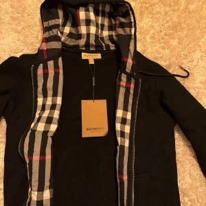 Svart Burberry kofta  - Svart kofta  från Burberry med klassiskt rutig insida på huvan och dragkedja framtill. Jackan har broderad Burberry-logga på bröstet och praktiska fickor. Perfekt för dig som vill ha en stilren och ikonisk look.