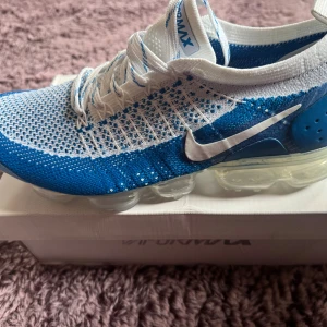 Nike Air Vapormax Flyknit 2 blå/vit, helt nya och oanvända. - Nike Air Vapormax Flyknit 2 i fräsch blå och vit färg. Skorna har stickad ovandel i Flyknit-material, genomskinlig platt sula med Vapormax-dämpning och klassisk vit Swoosh på sidan. Perfekt för dig som gillar sportig och modern stil.