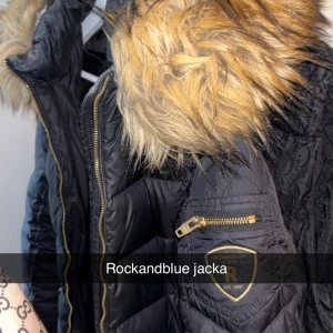 Rockandblue svart pufferjacka med päls - Svart pufferjacka från Rockandblue med stor fluffig päls på huvan och guldfärgade detaljer som dragkedja och logopatch på ärmen. Jackan har quiltad design och ger en riktigt schysst streetstil. Perfekt för kalla dagar när du vill se fresh ut.