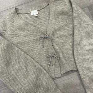 Beige stickad kofta med snörning - Mysig beige stickad kofta med v-ringning och långa ärmar. Koftan har två söta rosetter och är perfekt nu inför vintern 🫶🏻 obs passar även dig som har storlek S 
