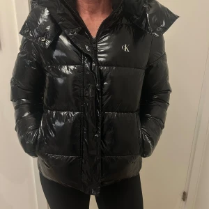 Svart pufferjacka från Calvin Klein - Snygg svart pufferjacka från Calvin Klein med glansig finish och logga på bröstet. Jackan har huva med snörning, dragkedja framtill och två fickor. Perfekt för kalla dagar och har en modern, oversized look. Den har inga defekter och är knappt andvänd❤️❤️