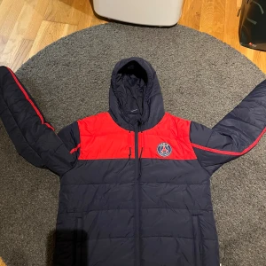 PSG pufferjacka med huva - Snygg pufferjacka från PSG i mörkblått och rött med klubbens emblem på bröstet. Jackan har huva med snörning, dragkedja framtill och sportig design med röda detaljer längs ärmarna. Perfekt för dig som älskar fotboll och vill ha en cool look.