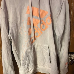 Ljusgrå hoodie från Adidas med logga - Ljusgrå hoodie från Adidas med stor aprikosfärgad logga på framsidan. Tröjan har huva med snörning, magficka och långa ärmar. Perfekt för chill eller träning, och den mjuka bomullen gör den riktigt skön att ha på sig.