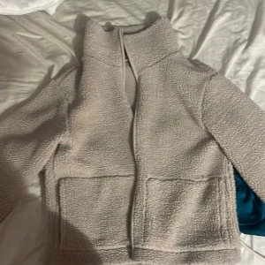 Beige teddyjacka med dragkedja - Mysig beige teddyjacka med hög krage och två stora fickor framtill. Jackan har långärmade ärmar och stängs med dragkedja. Perfekt för kalla dagar när du vill vara både chill och varm. Passformen är loose och materialet är fleece.