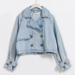 Gina jeansjacka - Säljer en ljusblå croppad jeansjacka med bred krage och dubbla rader bruna knappar framtill. Jackan har långa ärmar med snygga spänndetaljer vid mudden och en loose passform som ger en chill vibe. 
