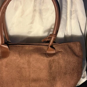 Brun axelväska i manchester och skinn - Snygg brun axelväska med mjuk manchester på utsidan och detaljer i brunt skinn. Väskan har rejäla handtag och en praktisk bältesdetalj med guldfärgat spänne. Perfekt storlek för att få plats med allt du behöver och stilren look.