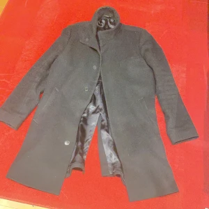 Svart rock från Tiger of Sweden - Snygg svart rock från Tiger of Sweden, modell EGAL. Jackan har klassisk rak passform, ståkrage och knäppning framtill. Yttermaterialet är en mix av ull och polyester, med ett slätt foder i viskos och polyester. Perfekt för dig som gillar stilrena och tidlösa plagg.