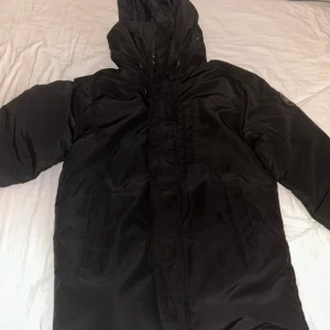 Canada Goose Jacka Black Label - Säljer en svart dunjacka från Canada Goose med stor huva och diskret logga på ärmen. Jackan har dragkedja och flera fickor framtill, perfekt för kalla dagar. Stilen är clean och jackan är långärmad med en rak passform.