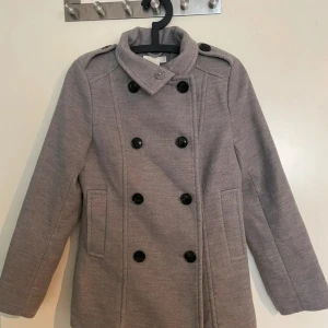Grå skepparkavaj från H&M, strl 36 - Snygg grå skepparkavaj från H&M med dubbelknäppning och svarta knappar. Jackan har axelklaffar, två fickor fram och klassisk krage. Perfekt för dig som gillar stilrena och tidlösa plagg med lite edge.