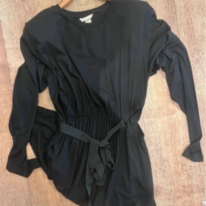Svart långärmad klänning med bälte - Snygg Nya svart klänning med långa ärmar och rund hals. Midjan är markerad med ett matchande tygbälte som knyts framtill. Klänningen har en enkel och stilren design, perfekt för dig som gillar minimalistiskt mode.