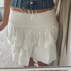 Vit volangkjol med broderade detaljer - Supersöt vit kortkjol med bred smock i midjan och volanger. Kjolen har broderade små blommor och är gjord i mjuk bomull, perfekt för varma dagar. Den har en romantisk vibe och passar dig som gillar detaljer och en feminin stil.