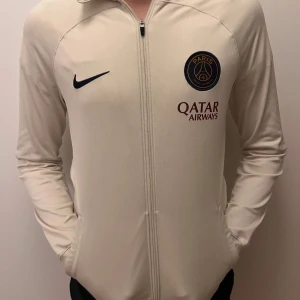 Beige PSG Nike zip-hoodie - Snygg beige zip-hoodie från Nike med Paris Saint-Germain-logga och Qatar Airways-tryck på bröstet. Jackan har huva, hel dragkedja och långa ärmar. Perfekt för dig som gillar fotboll och vill ha en sportig look.
