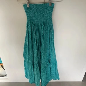 Turkos långkjol med mönster - Säljer en turkos långkjolmed geometriskt mönster och smockad överdel. Kjolen har en luftig och böljande kjol med volangdetaljer, perfekt för varma dagar. Materialet känns lätt och bekvämt mot huden.