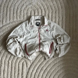 Vit vindjacka från Helly Hansen M - Snygg vit vindjacka från Helly Hansen med röda detaljer och broderad logga på bröstet. Jackan har dragkedja framtill och två fickor med röda kantband. Materialet är nylon med meshfoder och polyester. Perfekt för dig som gillar sportig och clean stil.