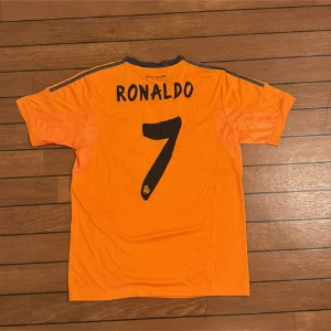 Real Madrid Ronaldo 7 orange Adidas tröja - Säljer en officiell Real Madrid fotbollströja från Adidas med Ronaldo och nummer 7 på ryggen. Tröjan är orange med mörkblå detaljer, korta ärmar och V-ringning. Materialet är lätt och andas, Climacool-teknologi. Fly Emirates-logga på bröstet.