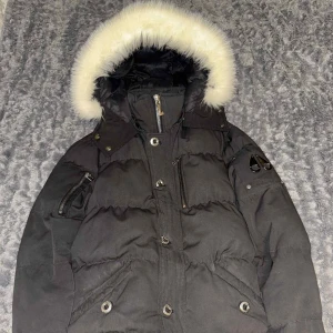 Svart Moose Knuckles vinter jacka med päls - Svart vinter jacka från Moose Knuckles med fluffig vit fuskpäls runt huvan. Jackan har flera fickor med dragkedja, knappar framtill och broderad patch på insidan. Perfekt för kalla dagar med sin tjocka och quiltade design.