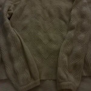 Beige stickad tröja med strukturmönster - Mysig beige stickad tröja med högt ribbat halsavslut och snyggt strukturmönster över hela plagget. Tröjan har långa ärmar och en lite loose passform, perfekt för lager på lager under kyliga dagar.