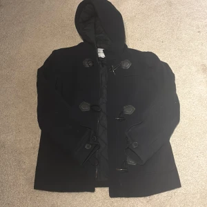 Svart skepparkavaj från Only & Sons - Svart skepparkavaj med huva från Only & Sons. Jackan har snygga knyt-detaljer framtill, stora fickor och quiltad insida. Perfekt för dig som gillar klassisk stil med modern touch. Passformen är normal och jackan är långärmad. Använd någon vecka under vintern är i helt ny skick har mest hängt där bara, väldigt varm och bra kvalité, köpt för cirka 1700kr så tänker att detta är ett bra pris för en nästan oanvänd jacka.