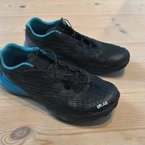 Svarta Salomon S/LAB sneakers - Salomon S/LAB sneakers i svart med blå detaljer och insida. Skorna har en sportig, aerodynamisk form och snörning med dragsko. Ovandelen är tillverkad i syntetmaterial med ventilerande mesh och platt sula för extra komfort. Perfekta för dig som gillar att köra swimrun