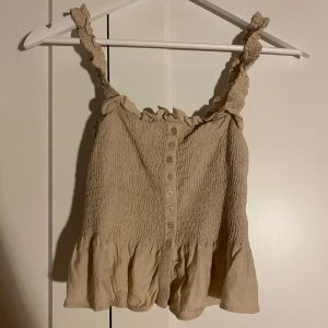 Beige linnetopp med volang - Supersöt beige linnetopp från H&M med smockad överdel, knappar framtill och volangkant nedtill. Axelbanden är rynkade och ger en gullig touch. Perfekt för varma dagar och enkel att matcha med jeans eller kjol.