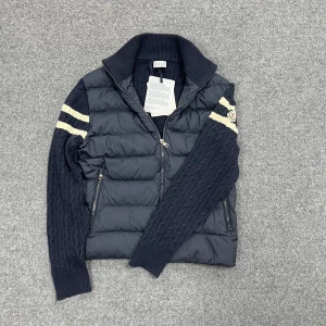 Mörkblå pufferjacka från Moncler - Snygg mörkblå pufferjacka från Moncler med stickade ärmar och två vita ränder på varje arm. Jackan har hög krage, dragkedja framtill och två sidofickor med dragkedja. Moncler-logga på vänster ärm. Kombinerar quiltad polyester med stickad ull för en unik look.