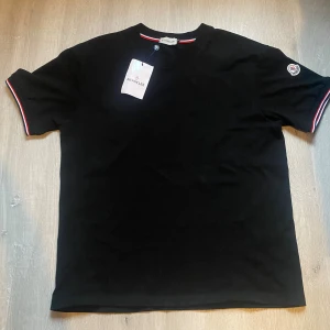 Svart Moncler t-shirt med logga - Svart t-shirt från Moncler i mjuk bomull. Klassisk rund hals och korta ärmar med snygga röd, vit och blå ränder vid ärmslut och hals. Moncler-logga på vänster ärm ger en exklusiv touch. Perfekt för dig som gillar stilrena plagg med detaljer.