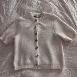 Vit kofta med rosettknappar från Zara - Superfin vit kofta från Zara med korta ärmar och söta silvriga rosettknappar framtill. Mjuk och mysig känsla. Ribbad kant vid hals, ärmslut och nederkant. Enkel och stilren design som passar till allt.