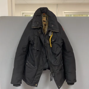 Svart Parajumpers dunjacka med päls - Svart dunjacka från Parajumpers med gul detalj vid kragen och flera fickor. Jackan har en stor huva med päls, ribbade muddar och dragkedja framtill. Perfekt för kalla dagar och har en cool patch på ärmen.             Rävsvansen till kragen kommer med jackan den sitter inte på nu för tillfället bara 