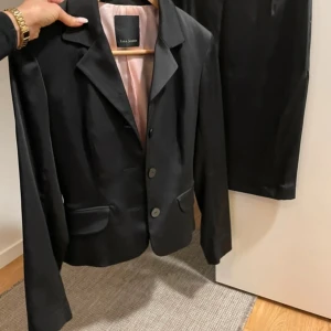 Svart kavaj och kjol från Tara Jarmon - Snygg svart kostym från Tara Jarmon med klassisk kavaj och tillhörande pennkjol. Materialet har en lätt glansig finish och ljusrosa foder. Perfekt för dig som gillar stilrena och eleganta looks.