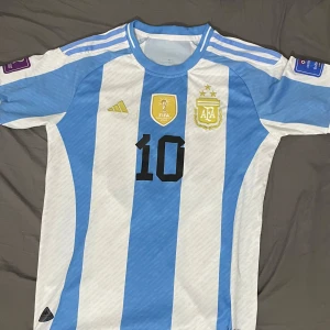 Argentina Messi #10 fotbollströja Adidas - Säljer en Argentina landslagströja med klassiska blå och vita ränder, tryckt nummer 10 och Messi på ryggen. Tröjan har korta ärmar, broderade märken och FIFA-emblem på bröstet. Perfekt för dig som älskar fotboll och Messi.
