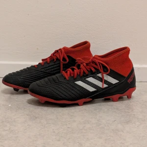 Adidas, Fotbollsskor firm ground- stl 41 1/2 Svart/Röd - Snygga Adidas Predator 18.3 FG fotbollsskor i svart och rött. Perfekt för fotbollsträningen eller matchen. Skorna har en strumpa som ger bra stöd runt ankeln och snörning för optimal passform. Använda ett fåtal gånger, haft dem som reservskor. Klassiska Adidas-ränder på sidan med Predator-logga på hälen. Perfekt för dig som vill ha grym kontroll på planen. Nypris: 1749 kr