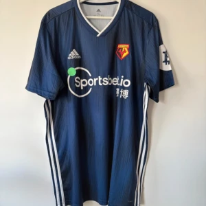 Watford FC blå Adidas fotbollströja XL - Snygg blå Watford FC fotbollströja från Adidas med korta ärmar och vitt V-ringad krage. Tröjan har diskret randigt mönster, klubbmärke på bröstet och Sportsbet.io sponsortryck. Klassiska Adidas-ränder längs sidorna. 