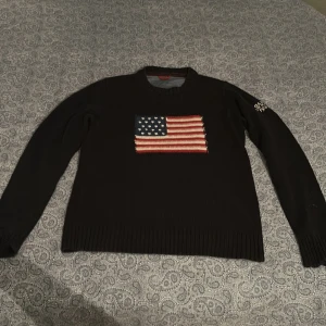 Blå stickad tröja med amerikansk flagga från Gant - Mörkblå stickad tröja från Gant med amerikansk flagga broderad på bröstet och texten 'GANT NEW HAVEN 1949' på ena ärmen. Klassisk rund halsringning och ribbade muddar vid ärmslut och nederkant. Den har en slitning på höger arm men annars är den i bra skick!