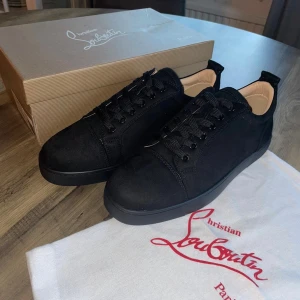 Christian Louboutin utan spikar - Snygga svarta sneakers från Christian Louboutin med klassisk röd sula och diskret logga. Skorna har rund tå, snörning och är tillverkade i mocka med detaljer i skinn. Perfekt för dig som vill ha en stilren och lyxig look med en ikonisk touch.