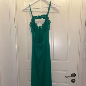 Grön långklänning med volang och snörning - Säljer en grön långklänning med smala axelband och snygg volangdetalj runt halsringningen. Klänningen har snörning framtill och en cool cut-out vid bröstet. Materialet känns lätt och flowy, perfekt för varma dagar. Passformen är normal och modellen är ärmlös.