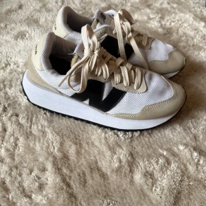 New Balance vita och beige sneakers  - Snygga sneakers från New Balance i vitt och beige med svart logga på sidan. Ovandelen är i mesh och mocka, med snörning och en platt, svart sula med vågigt mönster. Perfekta för dig som gillar stilrena och sportiga skor med retrovibe.