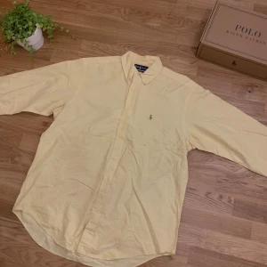 Gul skjorta från Ralph Lauren - Snygg ljusgul skjorta från Ralph Lauren med klassisk krage och broderad logga på bröstet. Modellen är Yarmouth och är gjord i 100% bomull oxford. Skjortan har knappar framtill och långa ärmar, perfekt för dig som gillar stilrena och tidlösa plagg. Har en liten fläck på ryggen samt ett namn inskrivet på inre kragen. (Se bild)