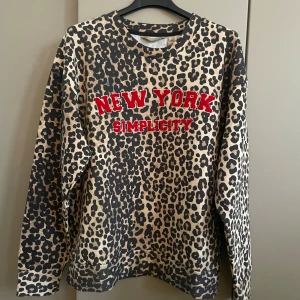Leopardmönstrad sweatshirt från Culture - Cool beige och svart leopardmönstrad sweatshirt från Culture med röd broderad text 'NEW YORK SIMPLICITY' på bröstet. Tröjan har rund hals, lång ärm och är tillverkad i mjuk bomull. Perfekt för dig som gillar statement-plagg och djurmönster.