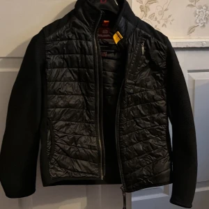 Svart quiltad jacka Parajumpers fin present till lillebror  - Svart jacka från Parajumpers med quiltad framsida och rygg, samt stickade ärmar och sidor. Jackan har dragkedja, hög krage och en gul detalj vid halsen. Märkespatch på ärmen och snygga kontraster mellan material. Perfekt för dig som gillar stilren och sportig look.