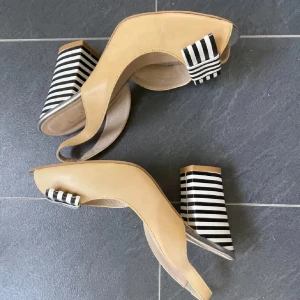 Beige pumps med randig klack och rosett - Unika pumps i beige skinn med öppen tå och bred klack i svartvitt randigt mönster. Framtill sitter en matchande randig rosett som ger extra attityd. Klacken är hög och stabil, perfekt för dig som gillar statement-detaljer och retrovibbar. Läder