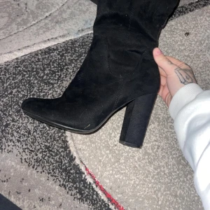 Svarta höga stövlar med klack - Säljer ett par svarta höga stövlar med bred klack och spetsig tå. Skorna är tillverkade i mocka och har en stilren, elegant look som passar till många outfits. Snygg siluett och platt sula framtill för extra komfort.