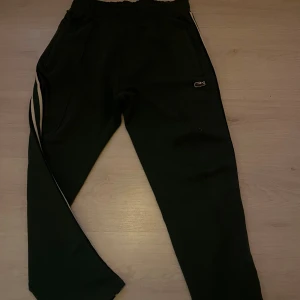 Svarta mjukisbyxor från Lacoste S - Svarta mjukisbyxor från Lacoste med klassisk krokodillogga på höften. Byxorna har elastisk midja, två sidofickor och vita ränder längs ena benet. Perfekta för chill eller träning, riktigt sköna och stilrena.