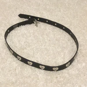 Svart bälte med hjärtan i silver - Svart bälte i skinn med coola silverfärgade hjärtan längs hela remmen och ett matchande silverspänne. Bältet är smalt och har en stilren look som passar till jeans eller kjol. Perfekt för dig som vill ha en edgy detalj med lite kärlek.❤️