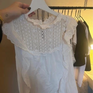 Vit spetsblus från MNG med volanger - Supersöt vit blus från MNG med spetsdetaljer och broderad framsida. Blusen har volanger längs axlarna och är ärmlös med rundad hals och knapp i nacken. Perfekt för dig som gillar romantisk och feminin stil.