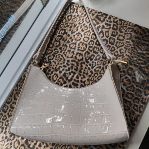 Vit hand/axelväska - Snygg vit axelväska i glansigt syntetmaterial med croco-mönster. Väskan har guldfärgade detaljer och dragkedja upptill. Perfekt storlek för det viktigaste och en stilren look.