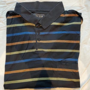 Randig pikéskjorta från Pierre Mâchén - Svart piké med ränder i blått, orange och gult från Pierre Hector. Den har klassisk krage, korta ärmar och tre knappar framtill. Materialet är mjuk bomull och det finns en liten bröstficka med broderad logga.